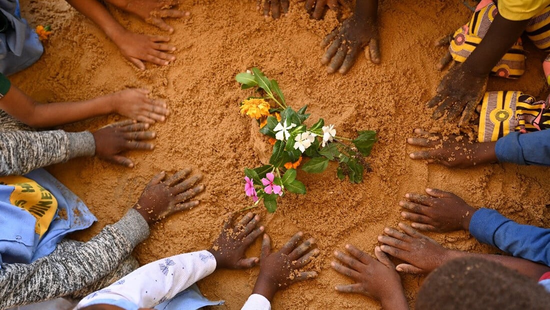 Kinderhände im Sand die einen Kreis um eine Blume bilden.