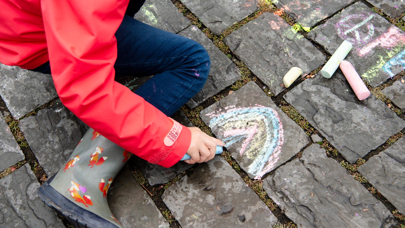 Kind malt am Boden einen Regenbogen mit Kreide.