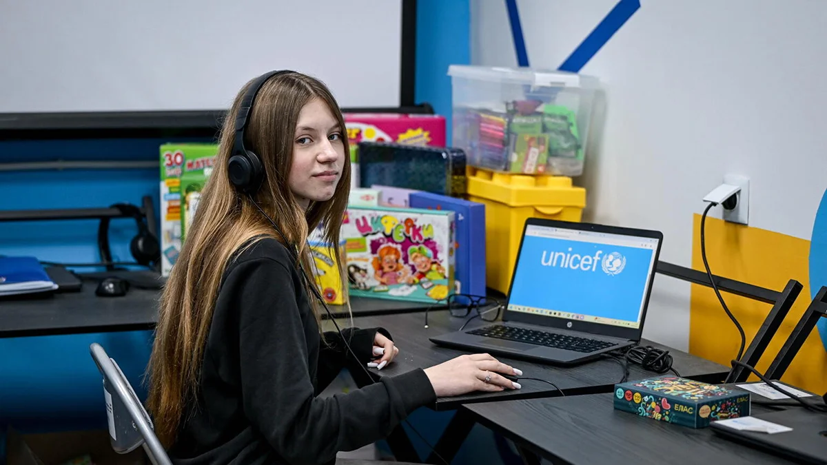 Ein Mädchen sitzt am Laptop mit Kopfhörern und schaut in die Kamera.