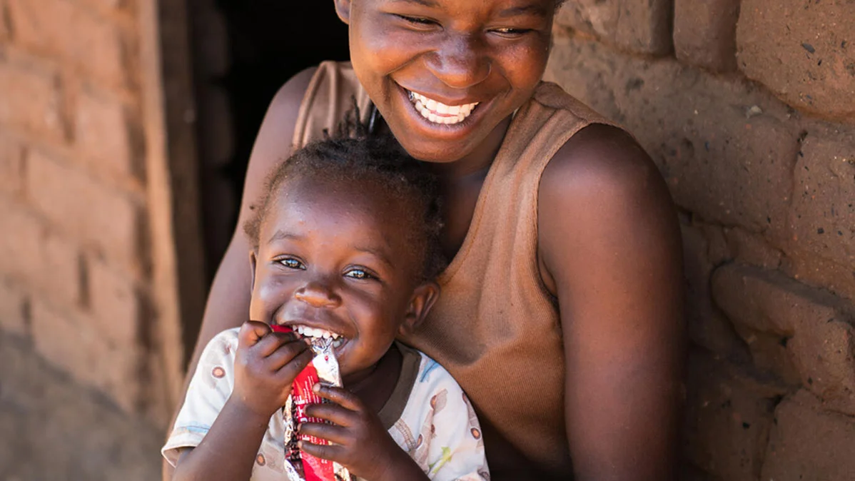 Die 20-jährige Edna Maunde lächelt, während ihre 2 Jahre und 4 Monate alte Tochter Christina Cosmas in ihrem Zuhause im Dorf Mavila (Zomba, Südmalawi) therapeutische Fertignahrung (RUTF) isst.