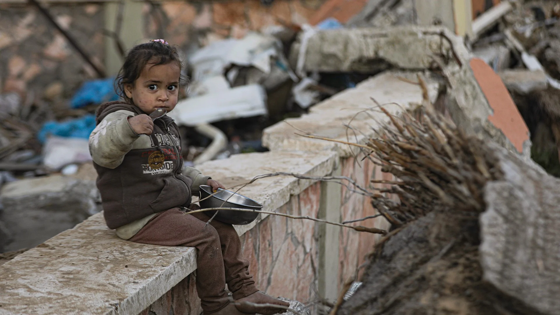 Kind sitzt auf Mauer mit Essen vor zerstoertem Hintergrund in Gaza