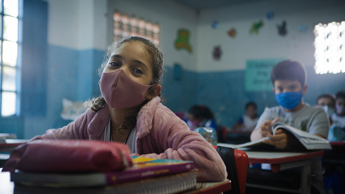 Kleines Mädchen sitzt mit Maske in einem Klassenzimmer.