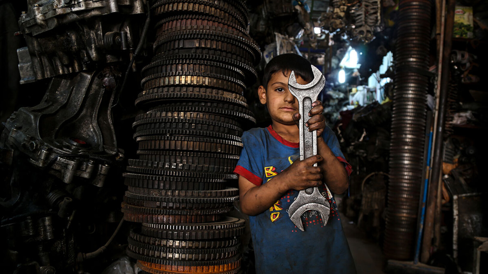 Ein kleiner Junge steht in einer Werkstatt und hält einen grossen Schraubenschlüssel in der Hand.