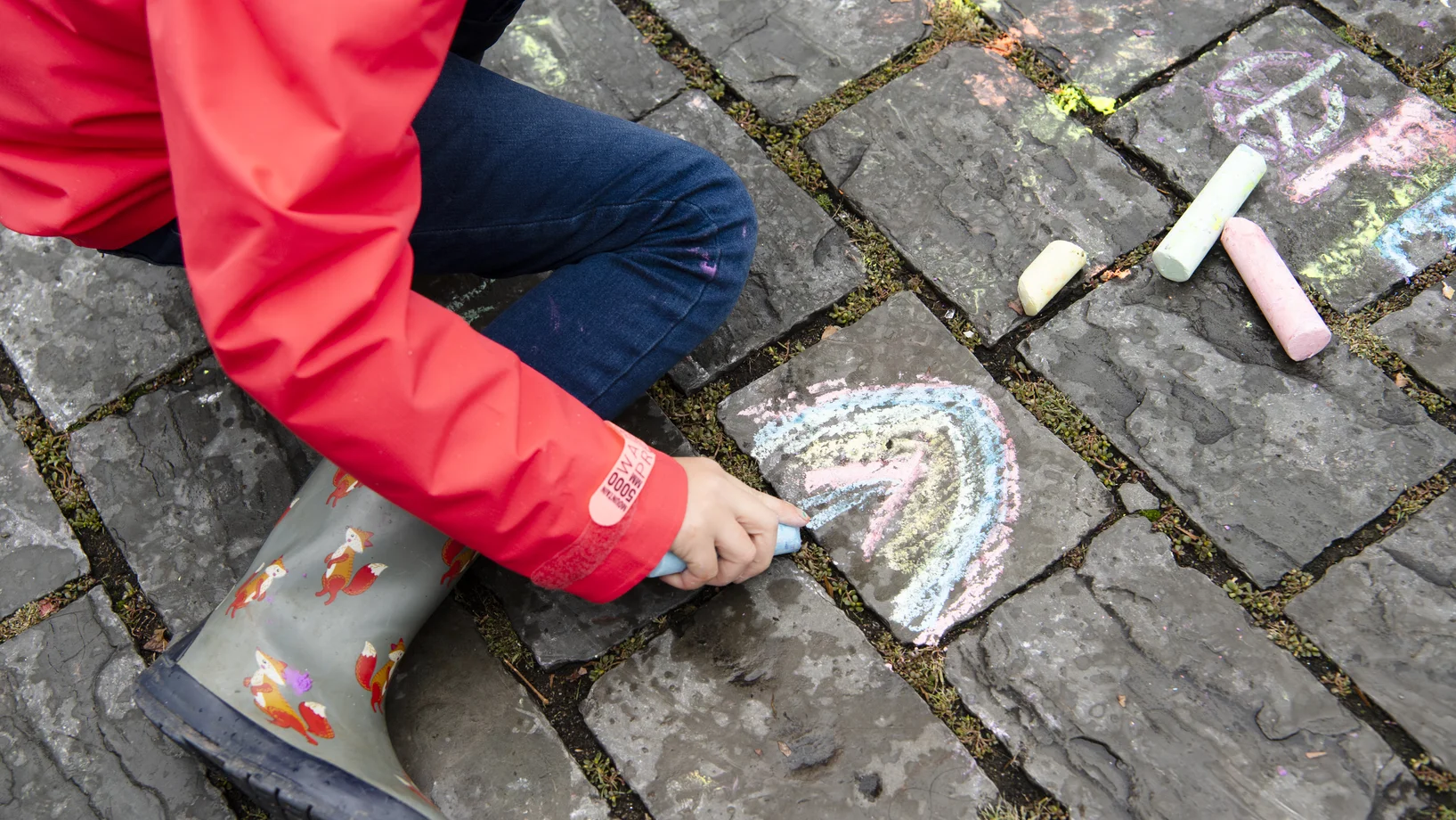Kind malt am Boden einen Regenbogen mit Kreide.