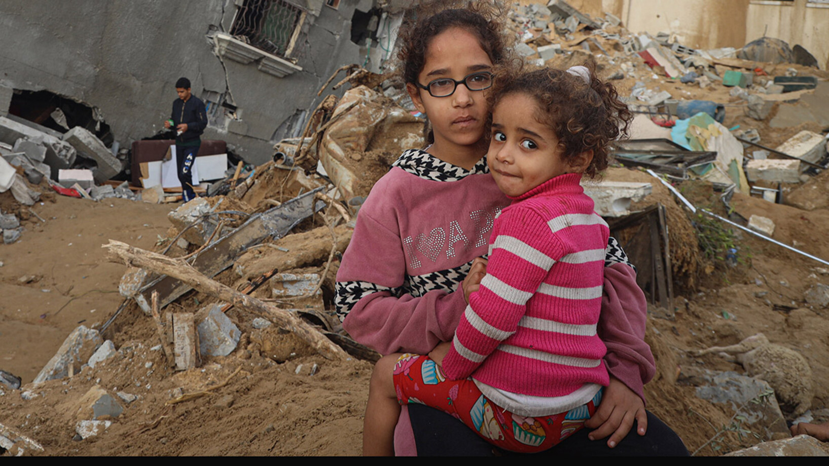 Ein Mädchen hät ihre kleine Schwester vor ihrem zerstörten Haus im Gazastreifen im Arm.