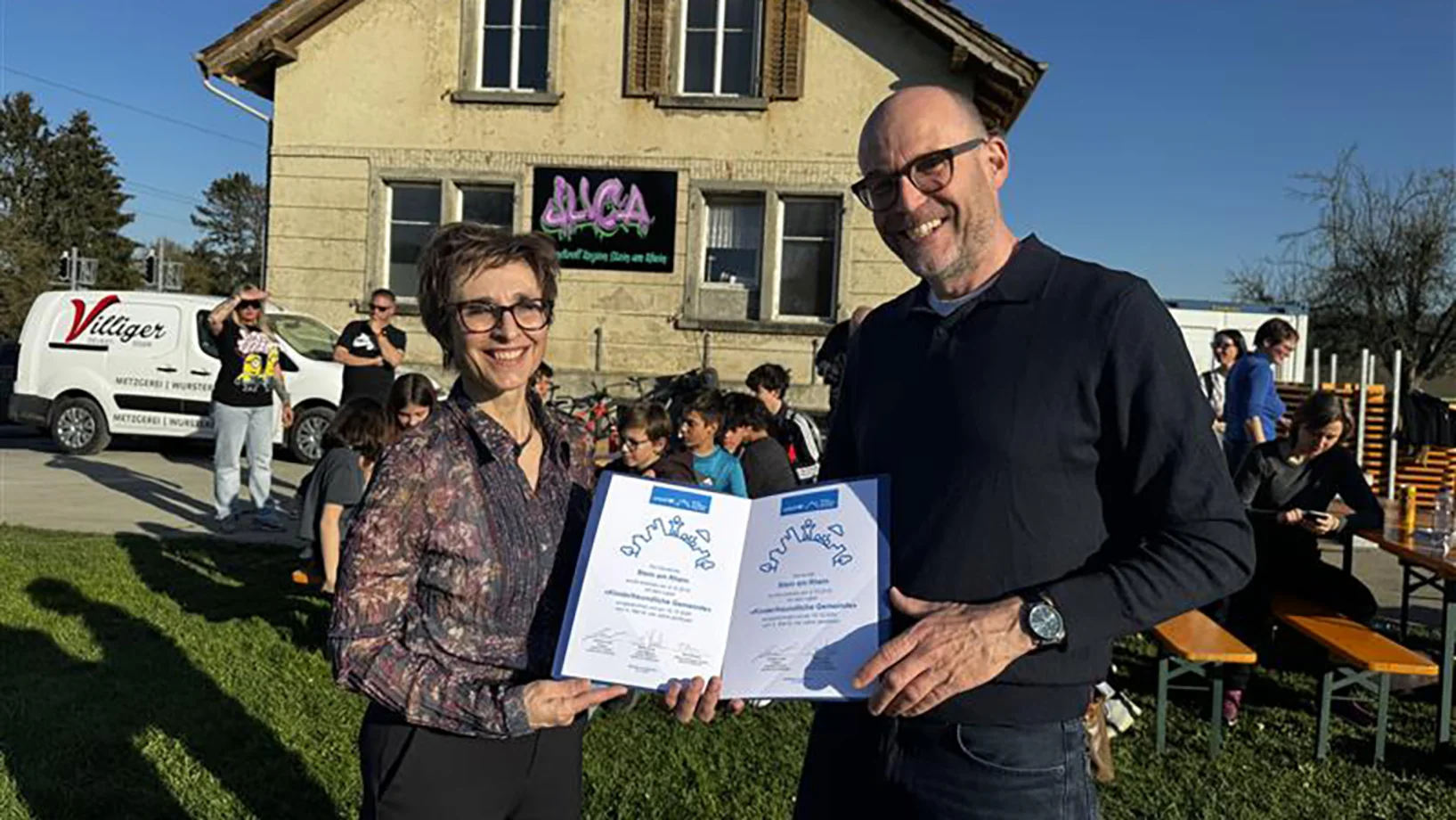 Zwei Menschen lächeln mit einer Urkunde in der Hand in die Kamera.