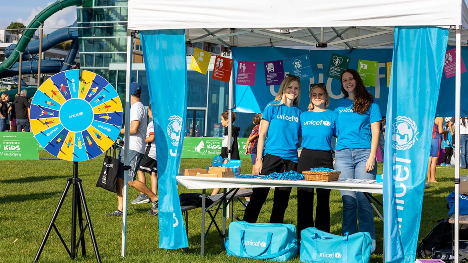 Drei Frauen stehen hinter einem UNICEF Stand und schauen in die Kamera
