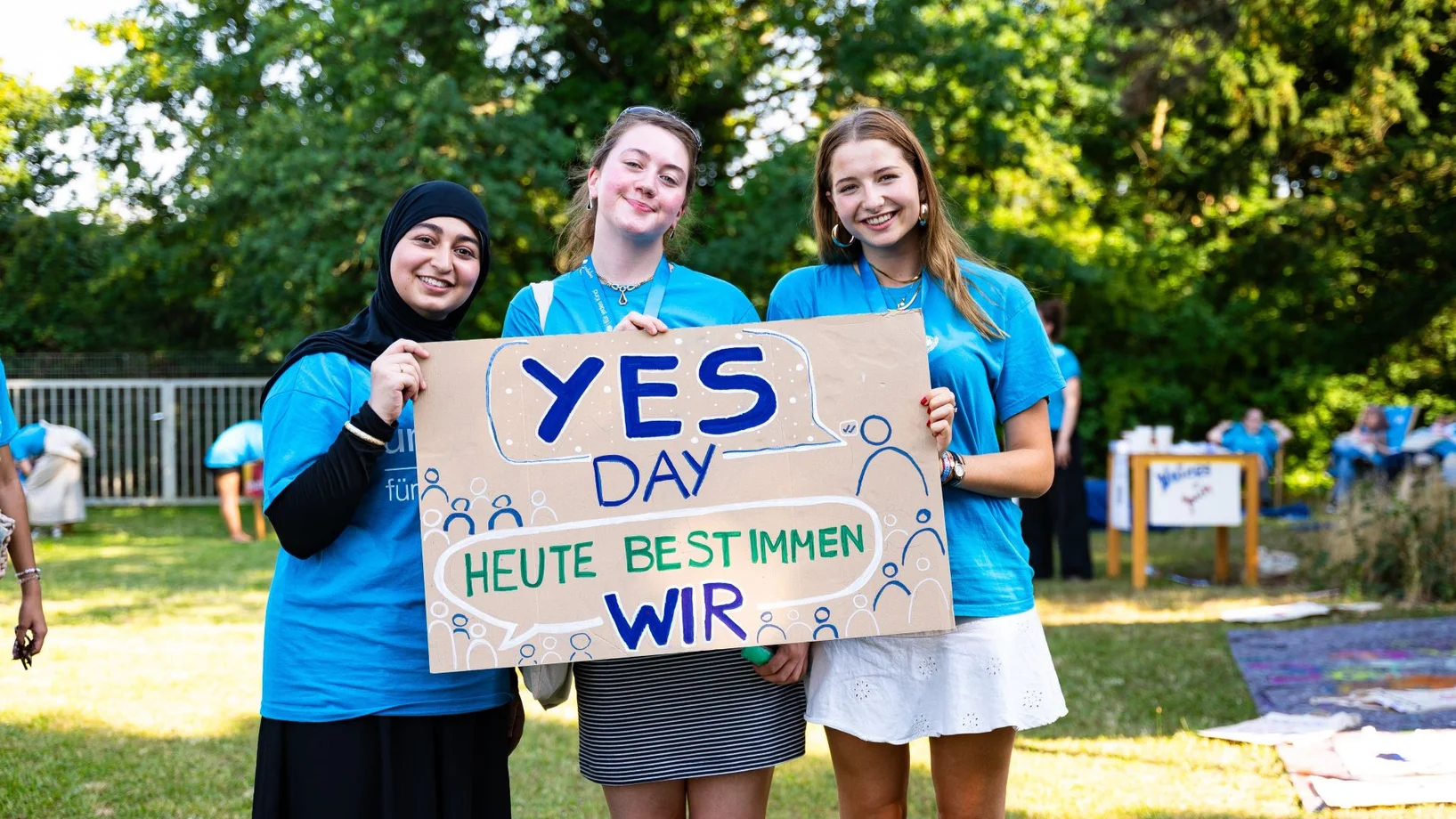 Drei Mädchen halten ein Schild mit dem Text "YES Day Heute bestimmen wir"