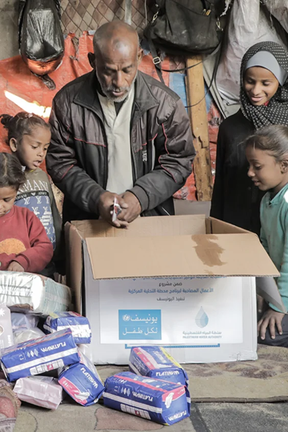 Ein Vater sitzt mit seinen Kindern in ihrer Notunterkunft vor einem UNICEF Karton, der mit Hygieneprodukten gefüllt ist.